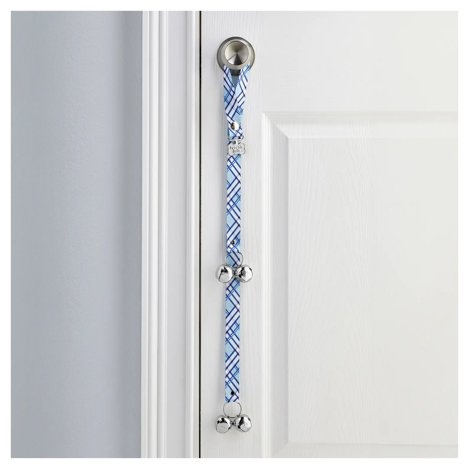Blue and white plaid dog doorbell strap hanging from a silver door handle on a white door, featuring a silver push-button at the top, a small bone-shaped PoochieBells tag near the center, and two sets of shiny metal jingle bells along the strap.