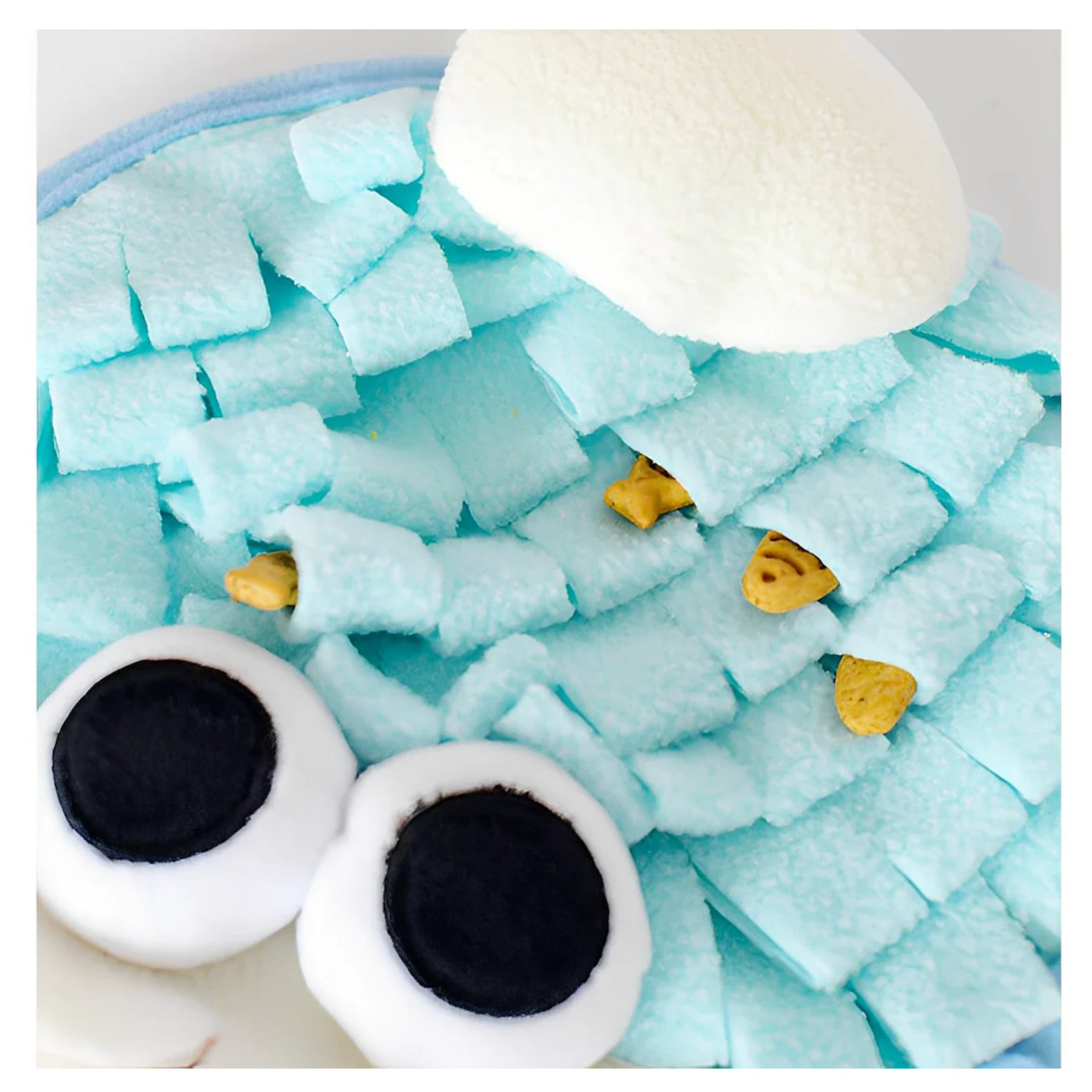 Close-up of the top portion of the ice-cream snuffle mat showing layered light blue fleece strips and a white plush topping, with small yellow treats tucked between the fabric folds and the toy’s large eyes partially visible.