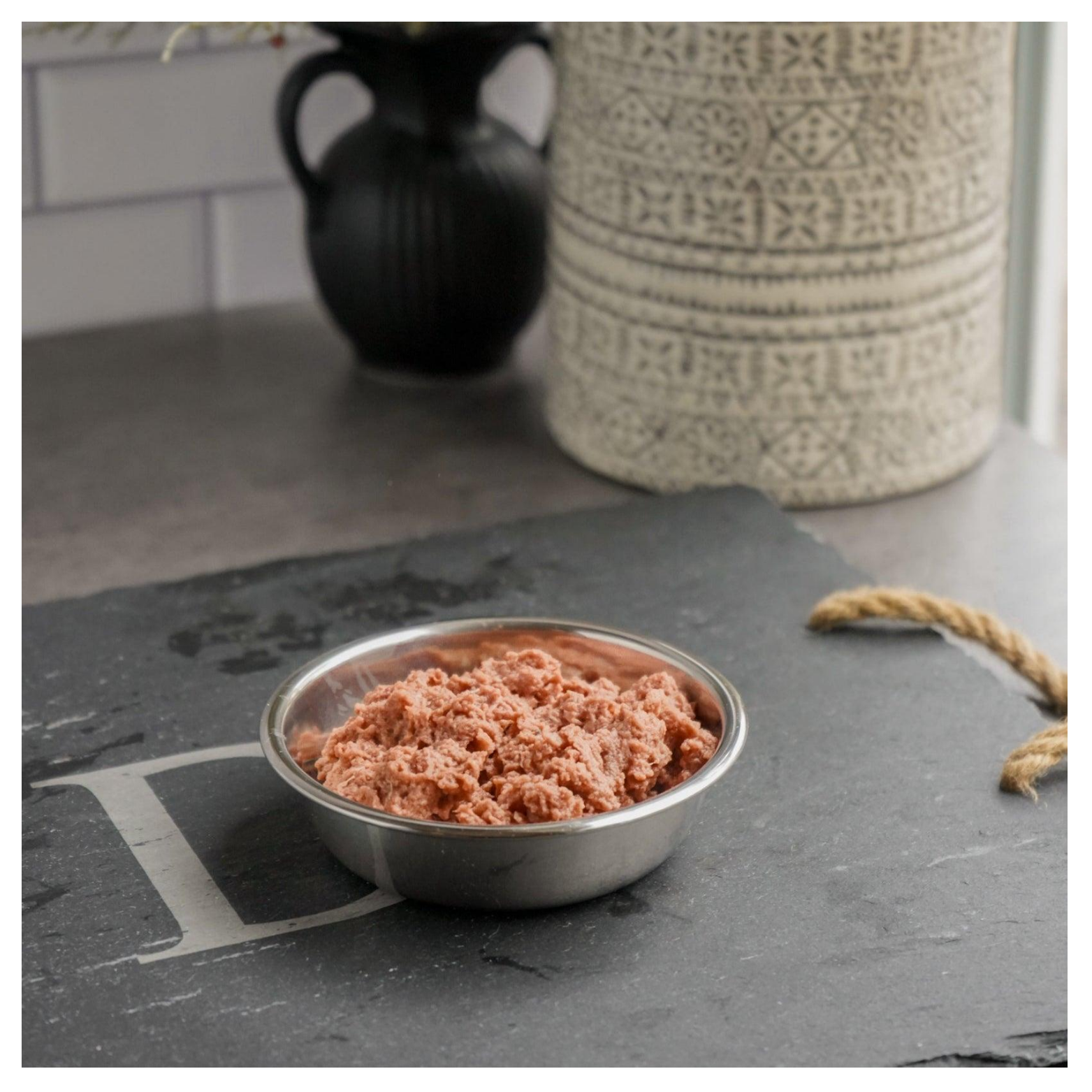 Metal pet bowl filled with ground raw quail meat sitting on a dark slate serving board in a kitchen setting.