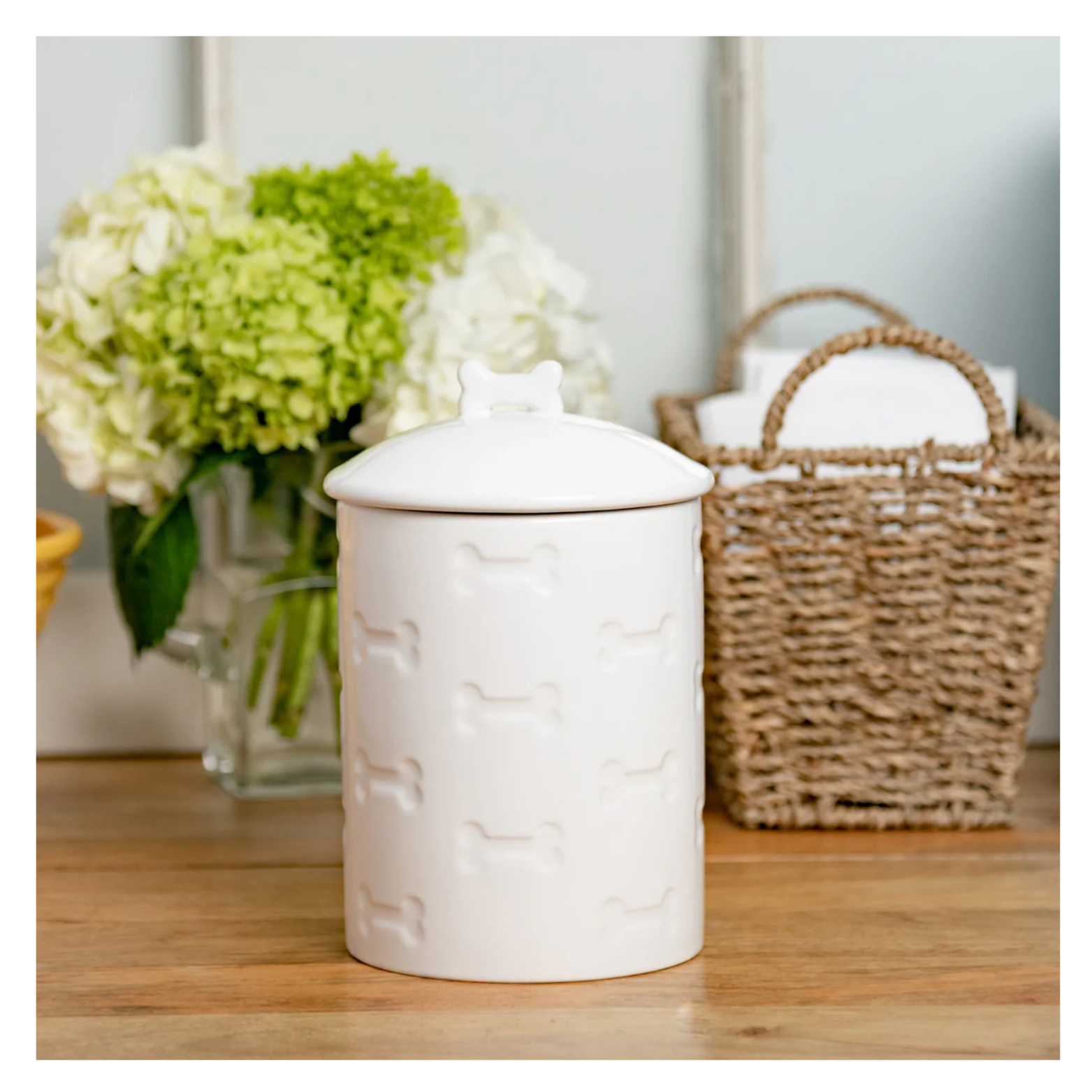 White ceramic pet treat jar with bone-shaped lid handle displayed on a wooden surface in a home setting, with flowers and a woven basket in the background.