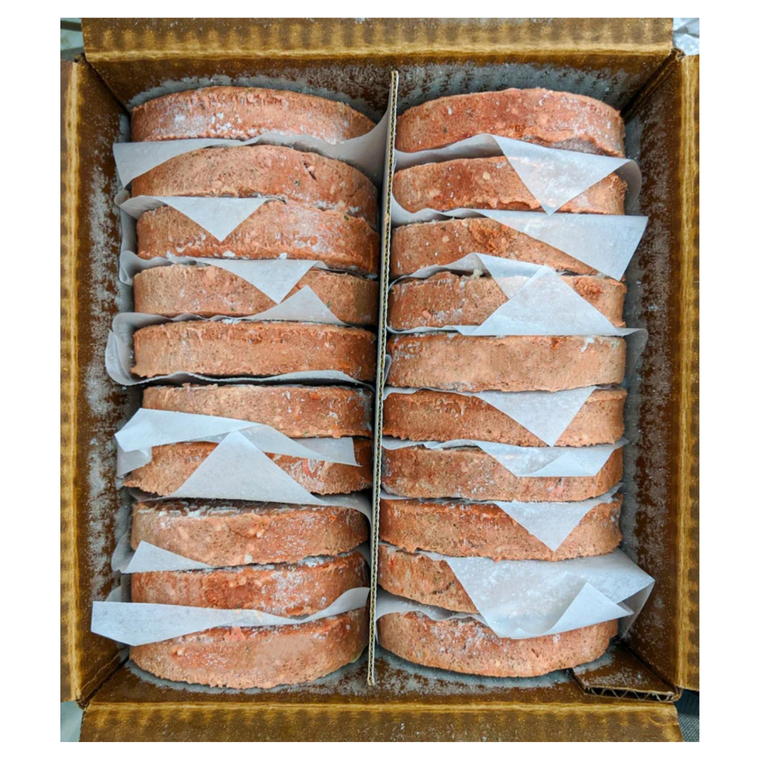 Top-down view of a cardboard box filled with frozen raw dog food patties stacked in two columns. The round patties are pinkish-brown, lightly frosted, and separated by sheets of white parchment paper.