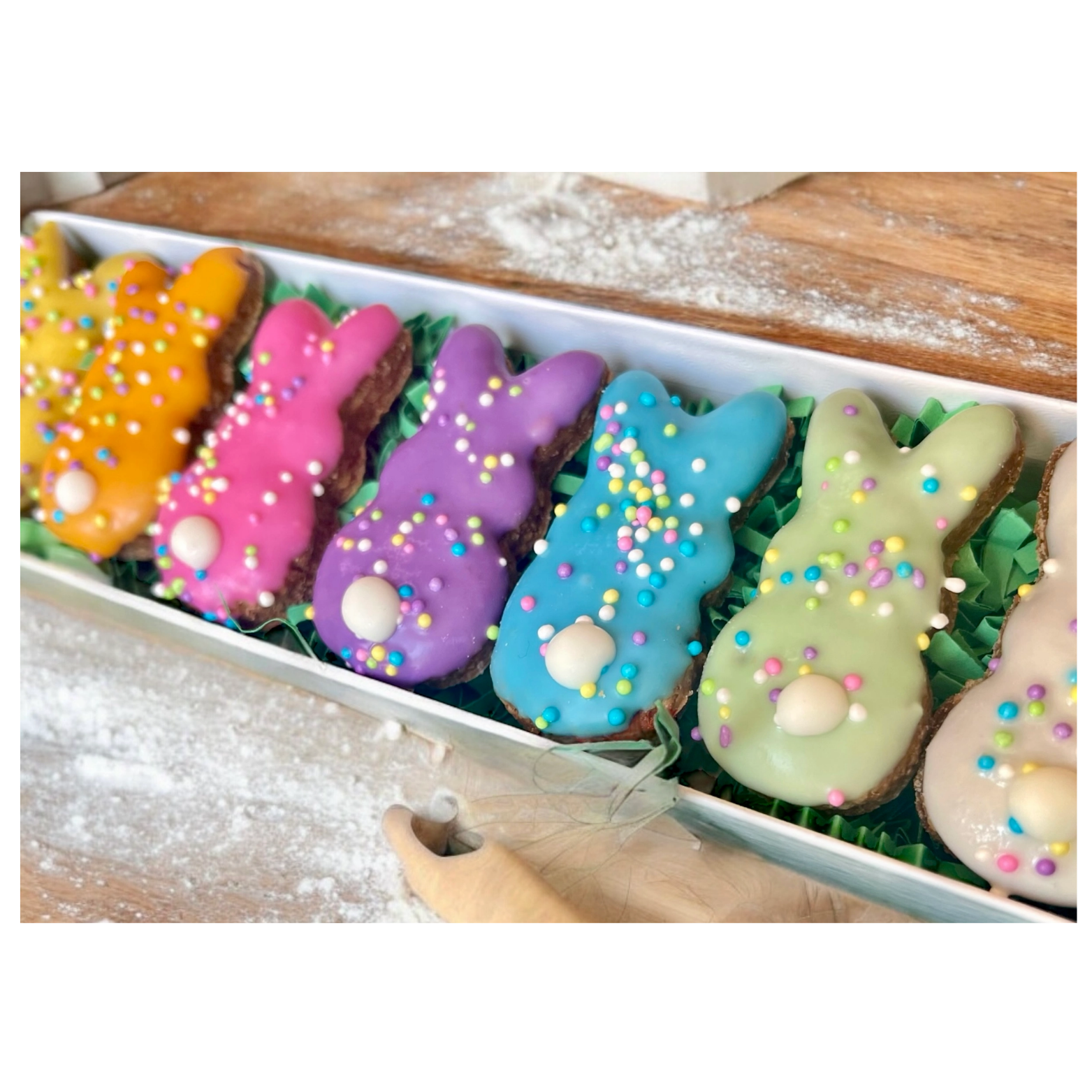 Close-up of a rectangular gift box filled with colorful bunny-shaped dog treats iced in pastel colors—yellow, pink, purple, blue, and green—topped with multicolored sprinkles and a white candy tail, resting on green paper filler with flour dusted on the wooden surface around it.