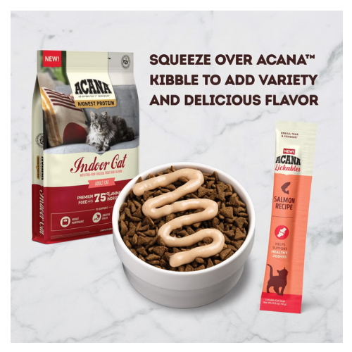 A bowl of dry cat kibble topped with a zigzag drizzle of creamy salmon lickable treat, shown beside an ACANA Indoor Cat kibble bag and a single ACANA Lickables salmon sachet on a marble surface.