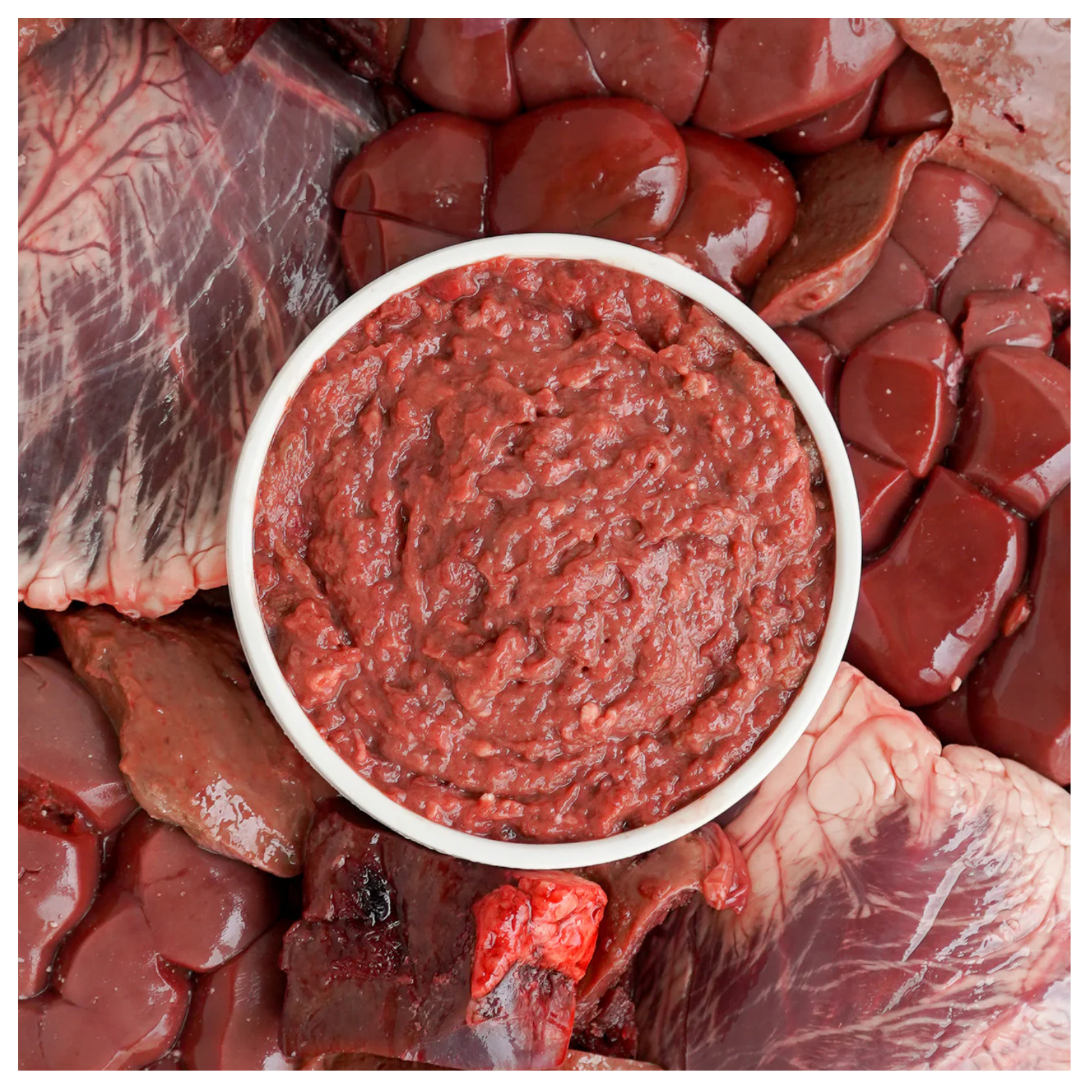 A bowl filled with ground beef organ mixture placed on a background of raw beef organs including liver and heart.