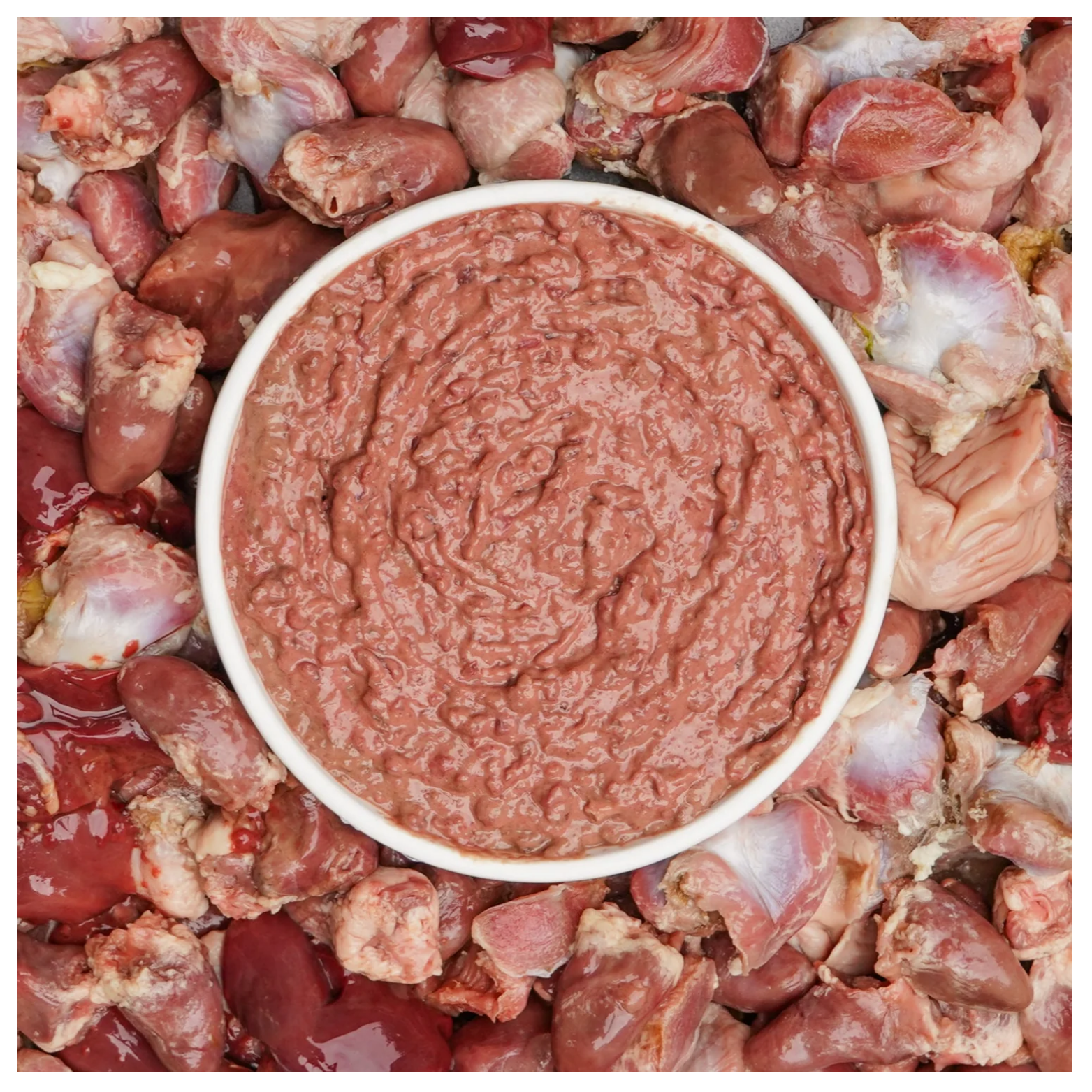 A bowl filled with ground turkey organ mixture placed on a background of raw turkey organs including hearts and gizzards.