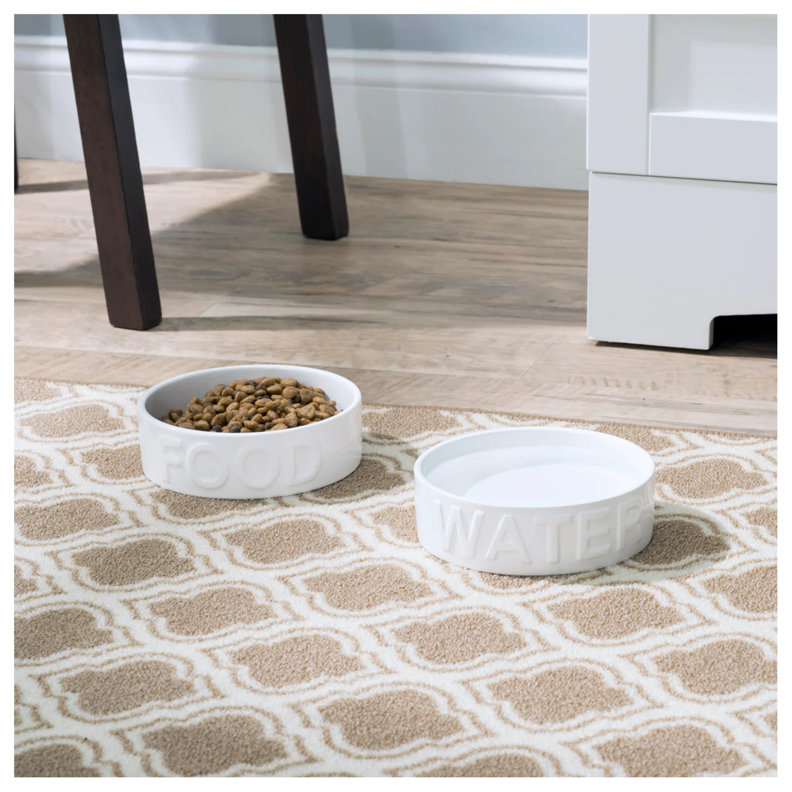White ceramic pet food and water bowls placed on a patterned rug in a home setting, with dry kibble in the food bowl and water in the water bowl.