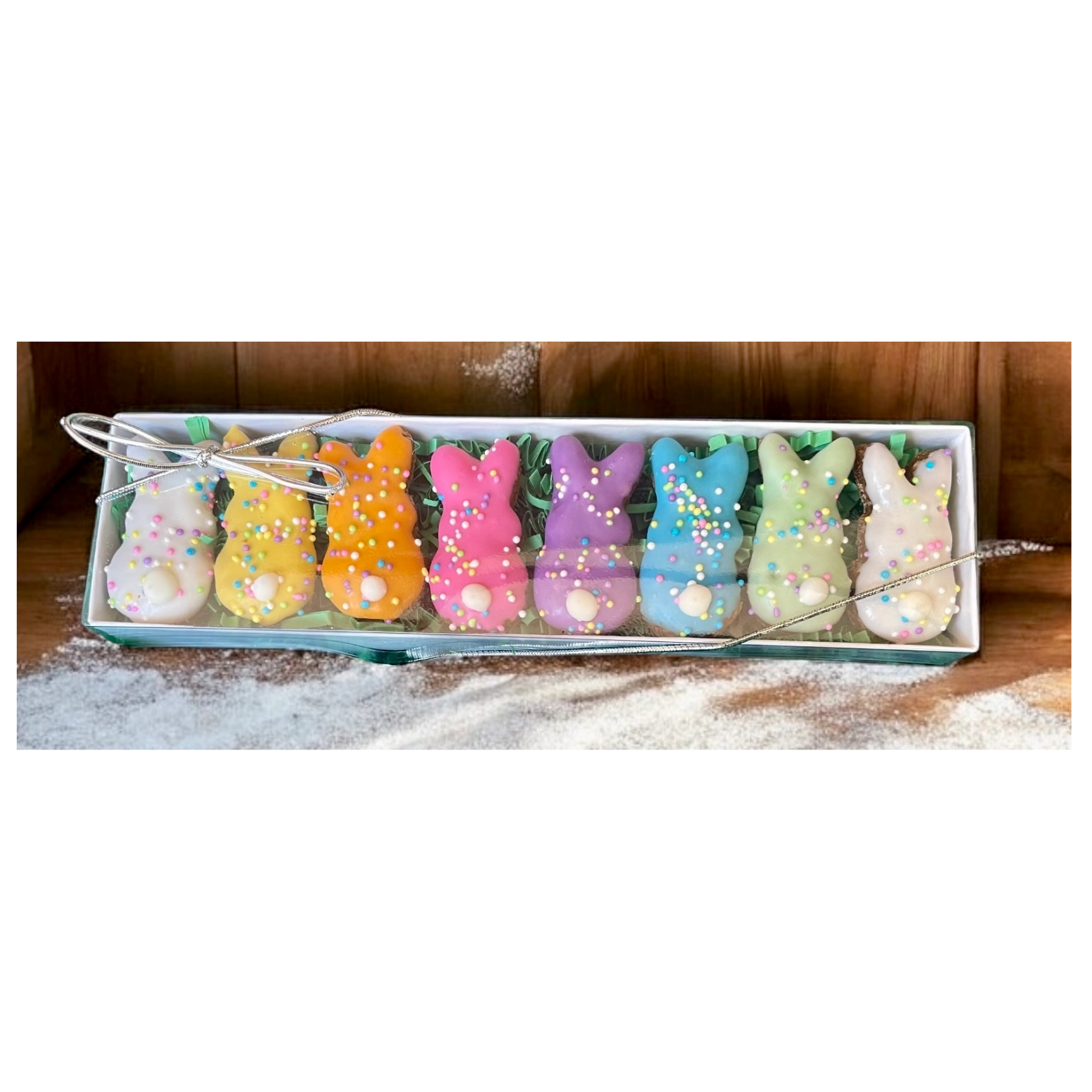 Long gift box of pastel bunny-shaped dog treats arranged in a row, each coated in bright icing and sprinkles, with a silver ribbon tied around the clear lid and powdered flour scattered on the wooden background.