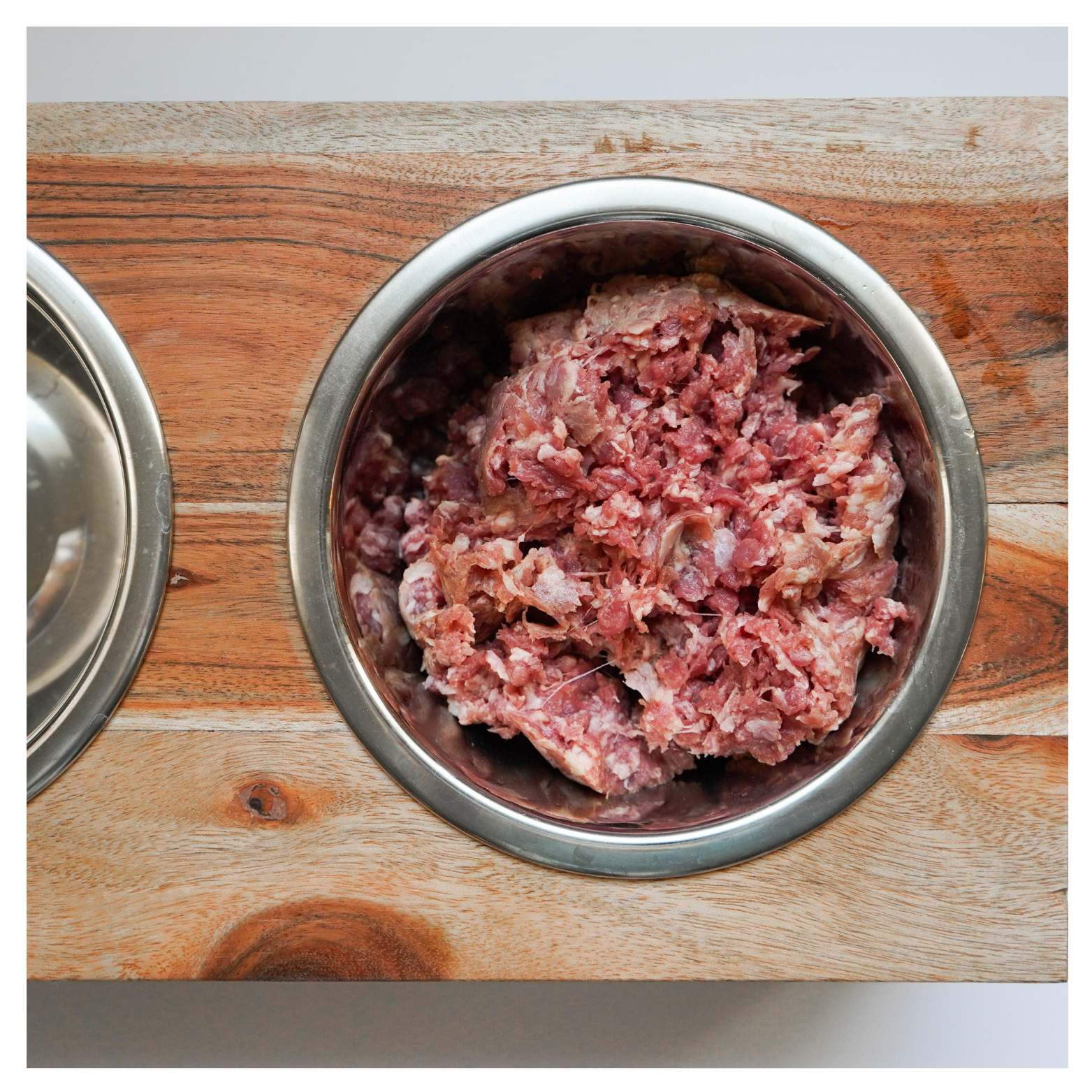 Top-down view of a stainless steel dog bowl filled with coarse ground raw quail meat set into a wooden feeder station.