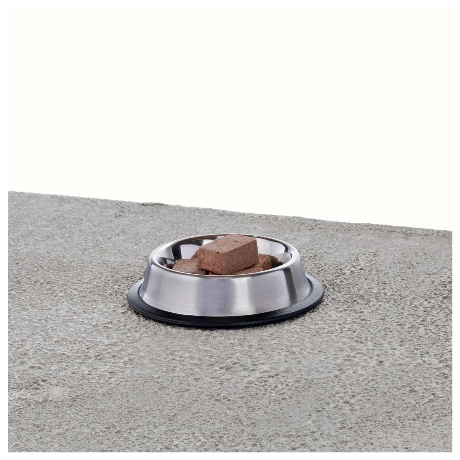 Stainless steel pet bowl containing several square frozen raw food nuggets on a textured gray surface.