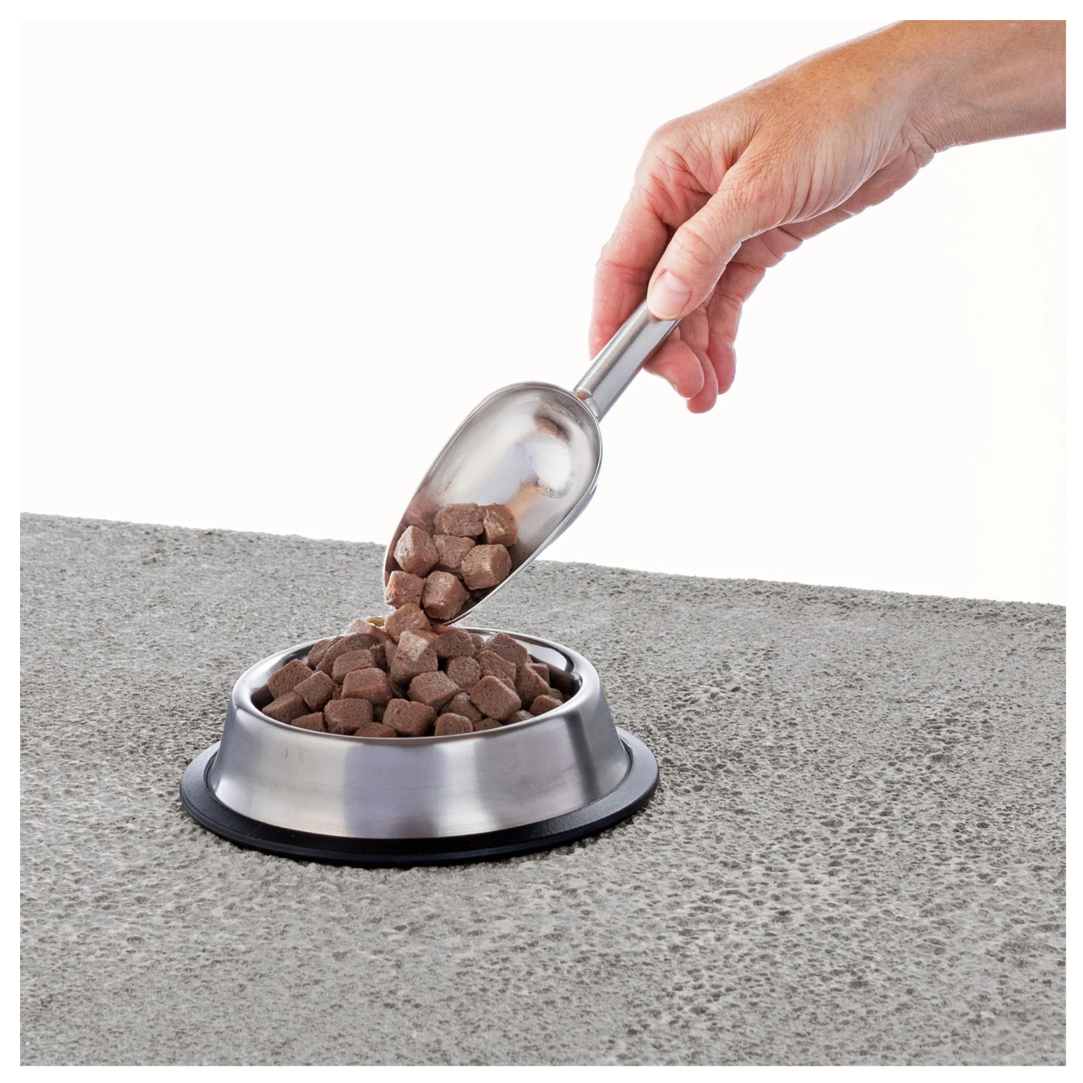 Hand holding a metal scoop pouring small cube raw food pieces into a stainless steel pet bowl placed on a textured gray surface.