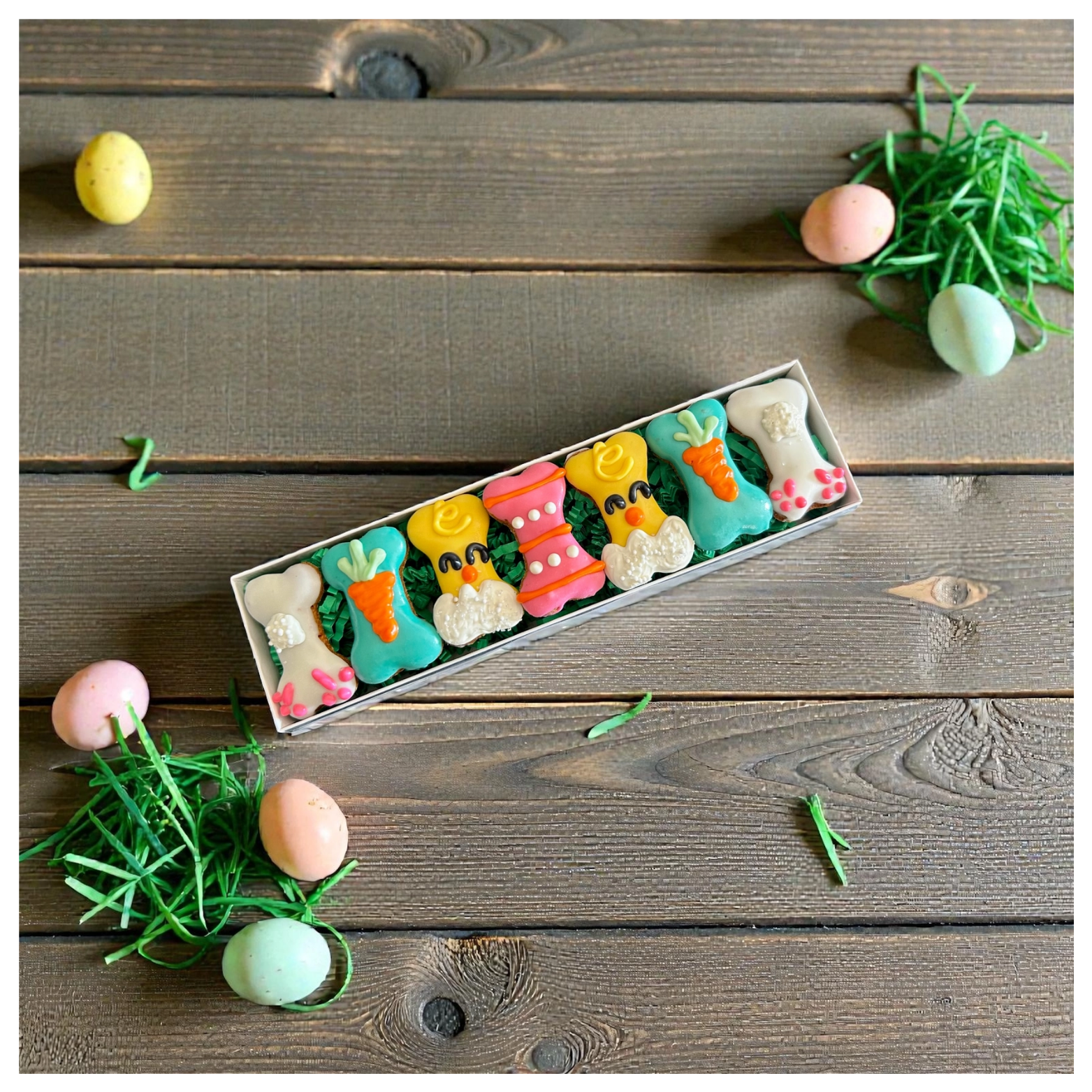 A top-down view of a rectangular box of Easter dog treats arranged in green filler on a rustic wooden table. Pastel eggs and green decorative grass are scattered around the box, emphasizing a festive holiday theme.