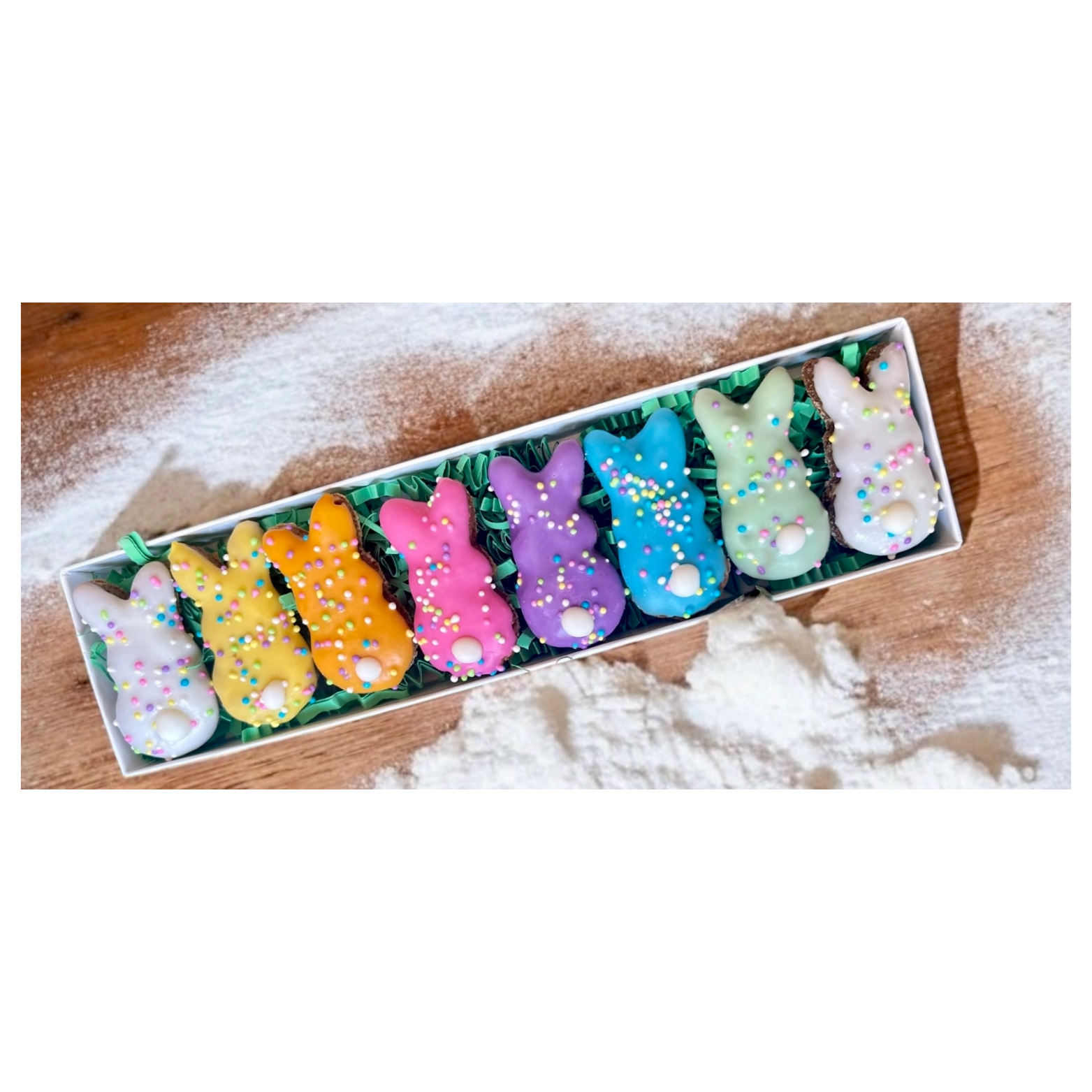 Top-down view of a narrow box containing rainbow-colored bunny-shaped dog treats with sprinkles and white tails, placed on green paper filler against a flour-dusted wooden surface.