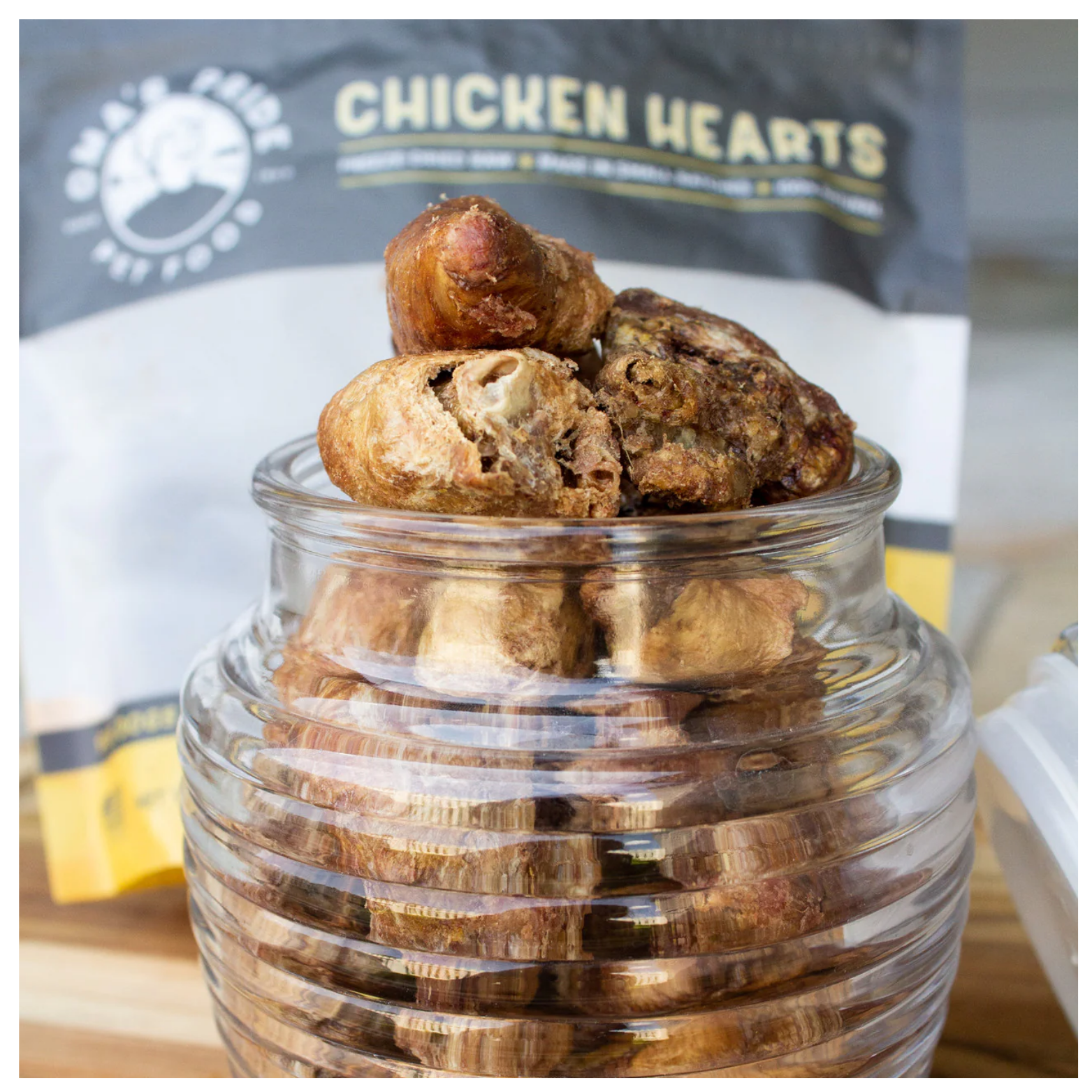 Glass jar filled with freeze-dried chicken heart treats on a wooden surface, with additional treats stacked on top and a blurred Oma’s Pride package visible in the background.