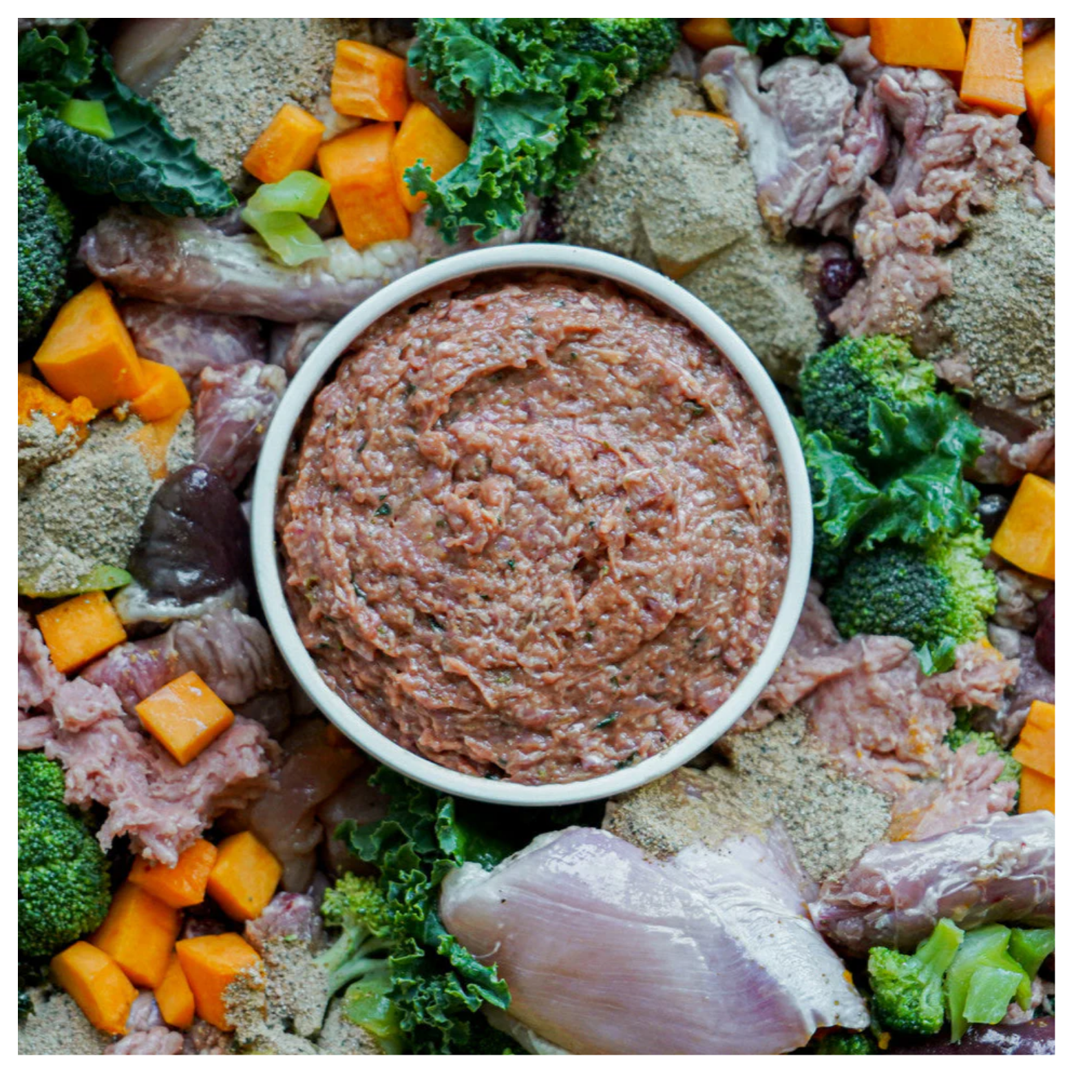 A bowl filled with ground chicken dog food mixture surrounded by raw chicken pieces, vegetables, and supplement cubes.